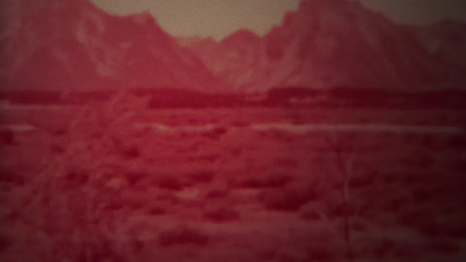 1967 Yellowstone, Wyoming 8mm frame showing a reddish, soft-focus view of low vegetation, open ground, and mountains under pale sky.