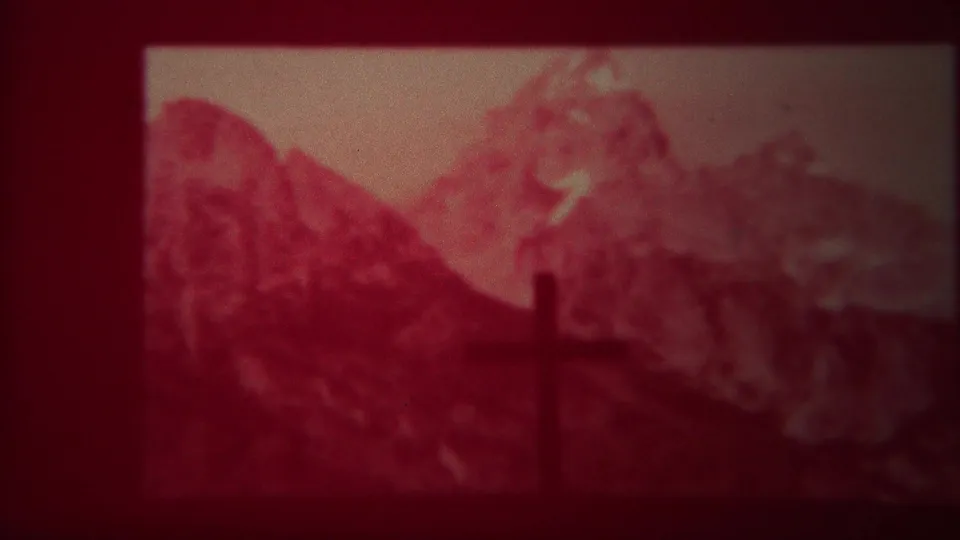 1967 Yellowstone home-movie frame with a dark cross-shaped foreground silhouette against red-tinted mountain peaks.