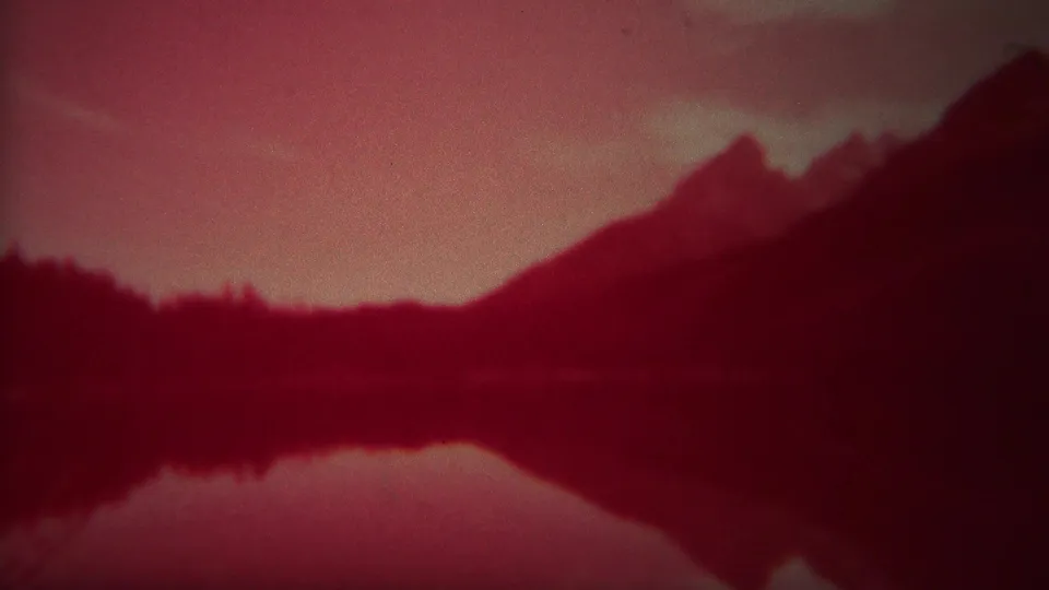 1967 Yellowstone home-movie frame of a dark shoreline and mountain reflected in still water under a red sky.