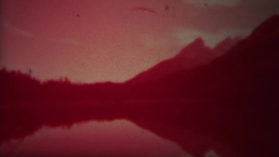 Yellowstone 1967 frame with a dark ridge, hazy sky, and mirrored lake surface in aged red-magenta 8mm color.
