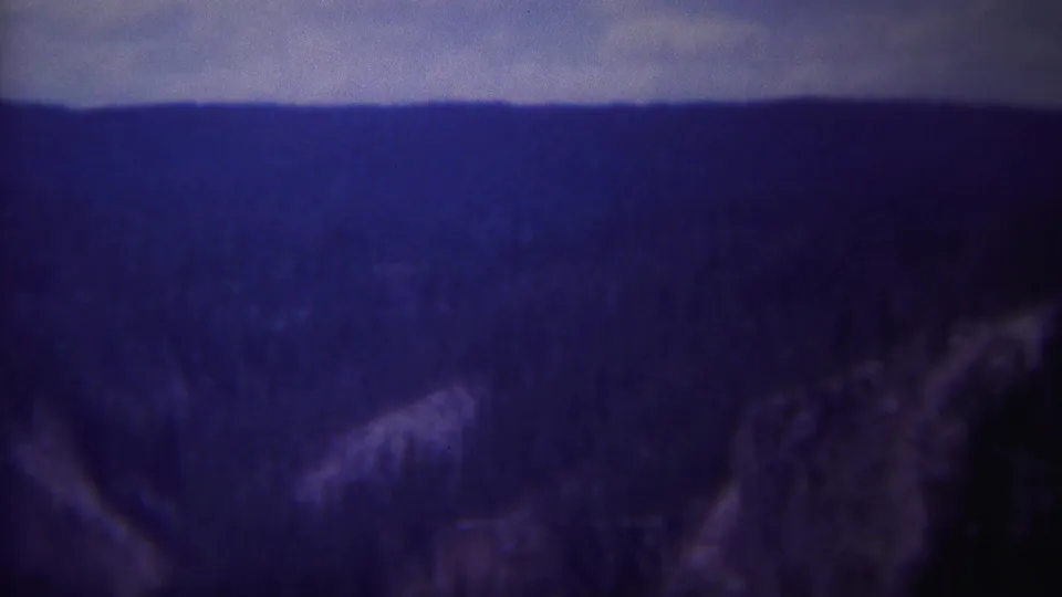1967 Yellowstone scenic frame with a blue-toned forested valley, distant ridge, and dark rocky foreground.