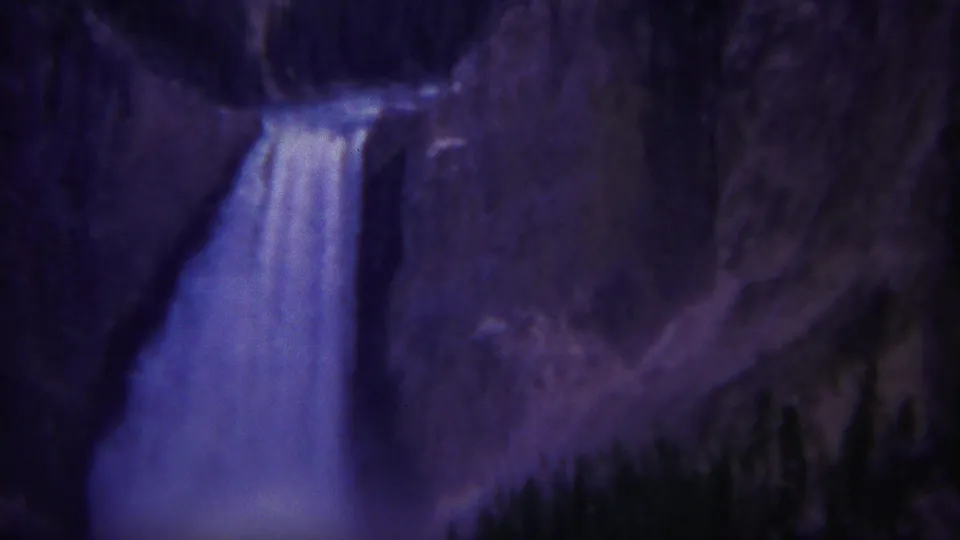 1967 Yellowstone home-movie still of a tall waterfall dropping beside steep rock and a dark evergreen tree line.