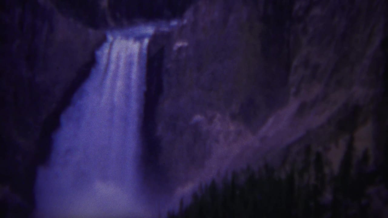 1967 Yellowstone home-movie frame showing a tall waterfall descending through a rocky cliff with dark conifers below.