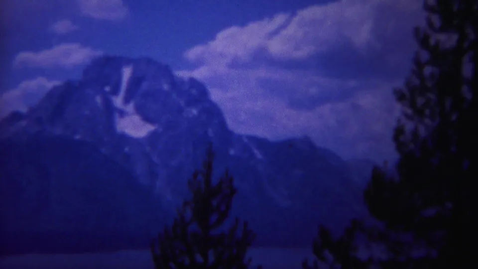 1967 Yellowstone home-movie view of a snow-marked mountain range beneath clouds, framed by dark pine silhouettes.
