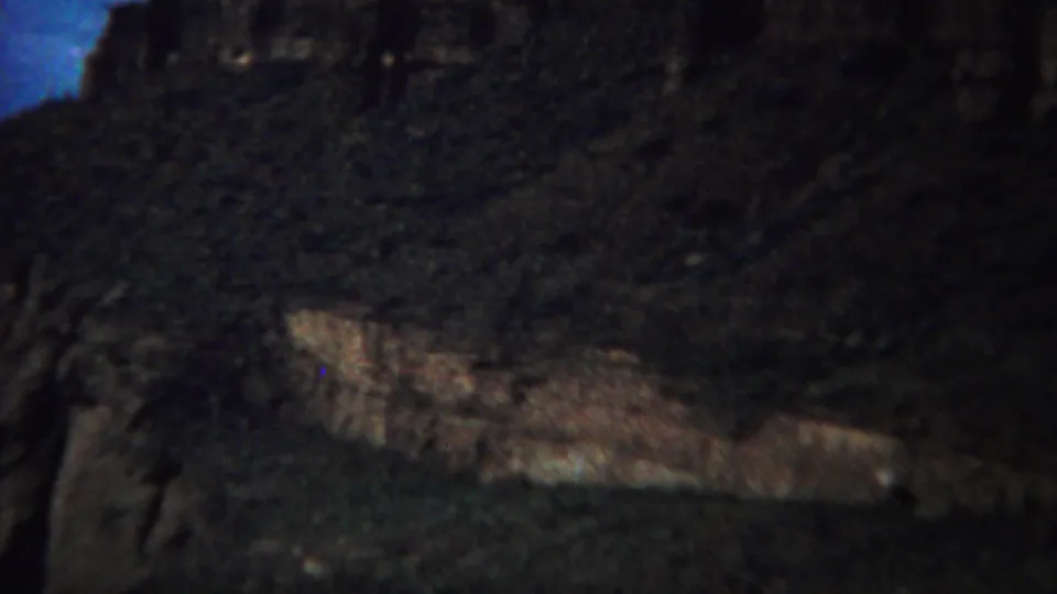 1967 Yellowstone frame showing a rugged canyon slope, pale ledge, dark vegetation, and blue sky at the edge of the frame.