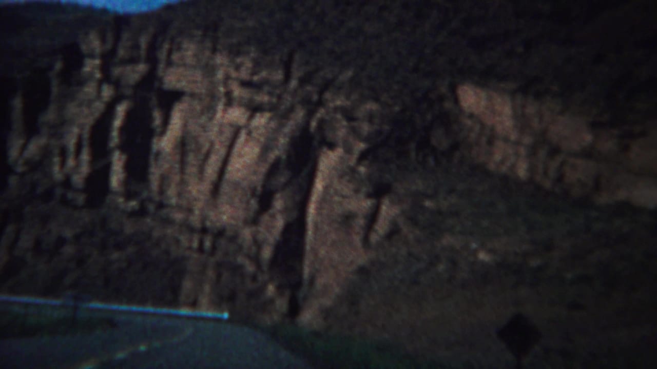Dark roadside view of a striated cliff face above a paved road and guardrail.