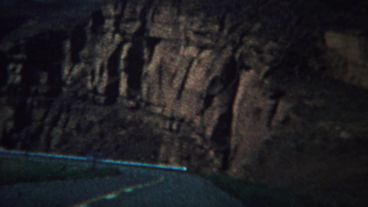 Tall rock wall with vertical striations seen from across a dark road.
