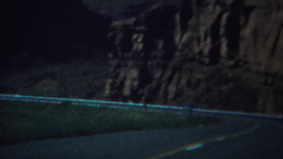 A dim 1967 Yellowstone travel frame shows a rugged cliff behind a roadside barrier and asphalt foreground.