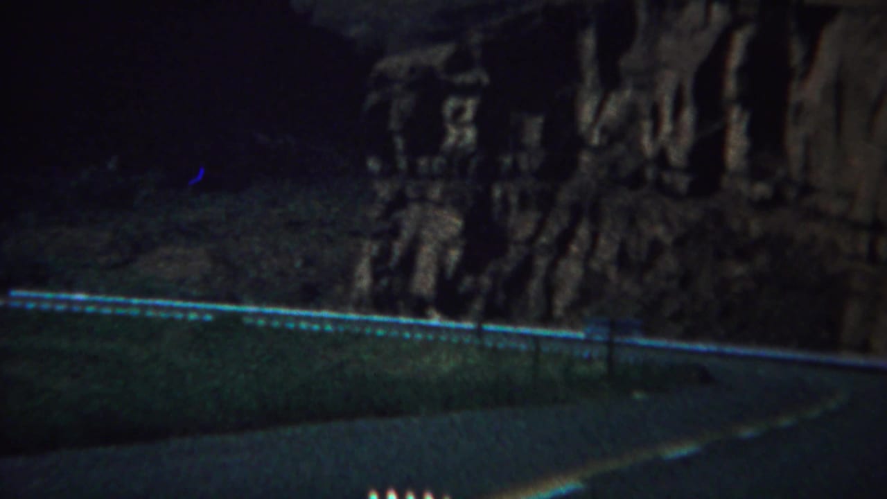 Shadowed mountain road curving below rocky cliffs and a pale guardrail.