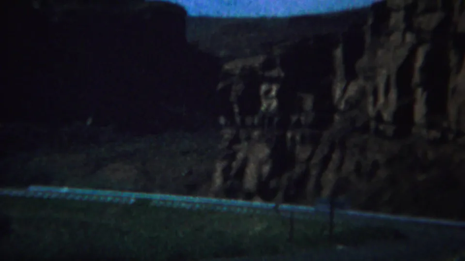1967 Yellowstone-area travel frame with a road edge, guardrail, and steep rock formations under low exposure.