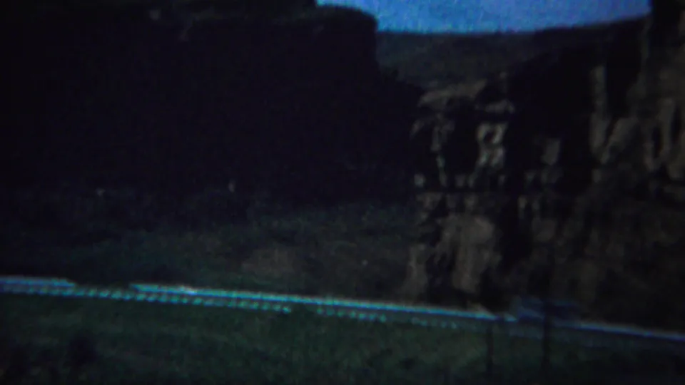Low-light 8mm Yellowstone-area frame from a moving vehicle, with guardrail, road edge, and shadowed rock slope.