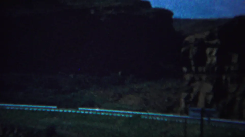 1967 Yellowstone-area 8mm frame showing a curving guardrail, dark roadway edge, and shadowed canyon rock.