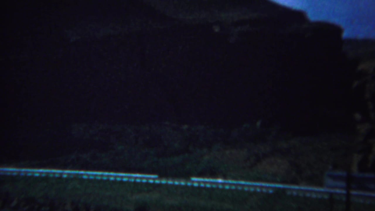 Dark roadside landscape with a pale road line and shadowed hills in the background.
