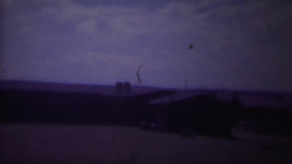 A 1967 8mm frame shows shadowed rural buildings beneath a cloudy sky, with a small dark mark near the upper right.