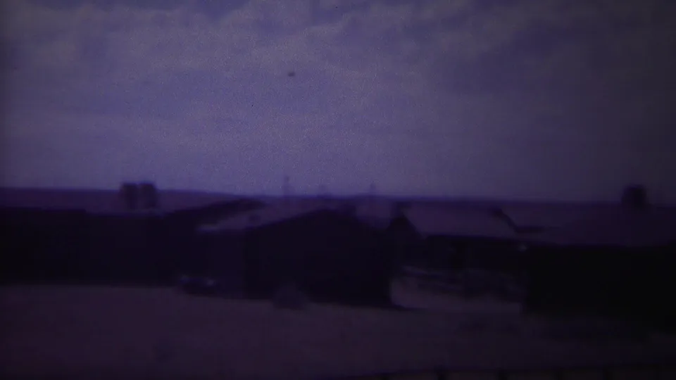 1967 Yellowstone, Wyoming home-movie frame showing low wooden buildings and dark roofs under a faded blue sky.