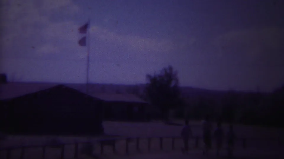 Distant figures walk through a faded 1967 Yellowstone, Wyoming town scene with low buildings, flags, trees, and a fence.