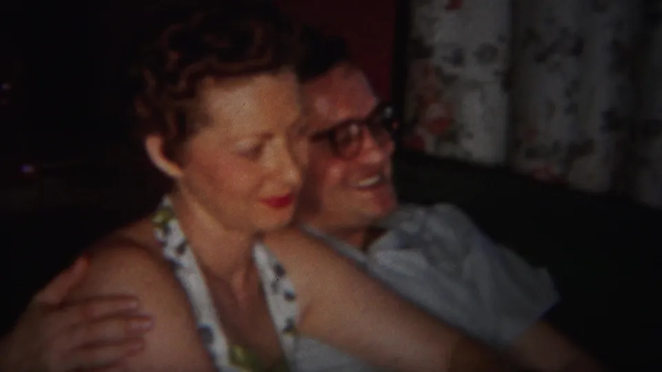 A close home-movie frame shows the woman seated forward while the man smiles behind her in the Westville interior.
