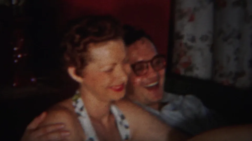 The couple smiles and leans together on a couch in the 1951 Westville home-movie scene, with floral curtains behind them.