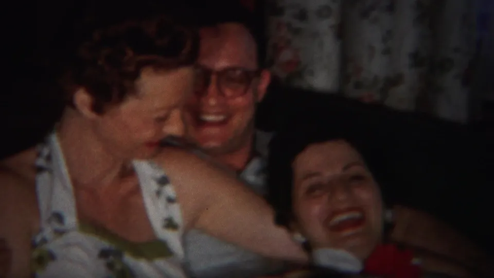 A woman leans toward a smiling man with glasses while another woman laughs beside them in the 1951 home interior.