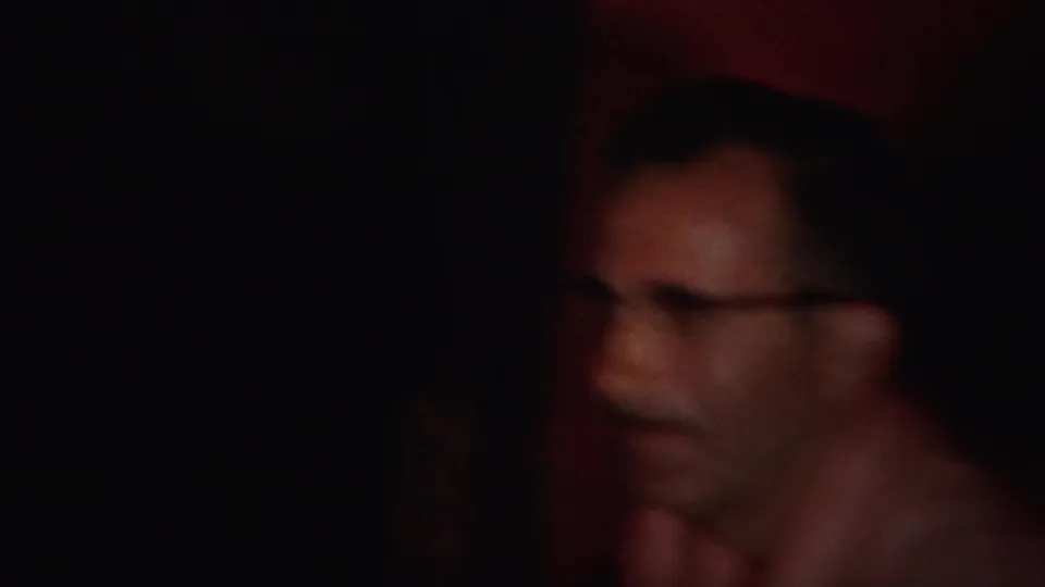 A man wearing glasses appears partially shadowed in the dim red-walled interior of the Westville home-movie scene.