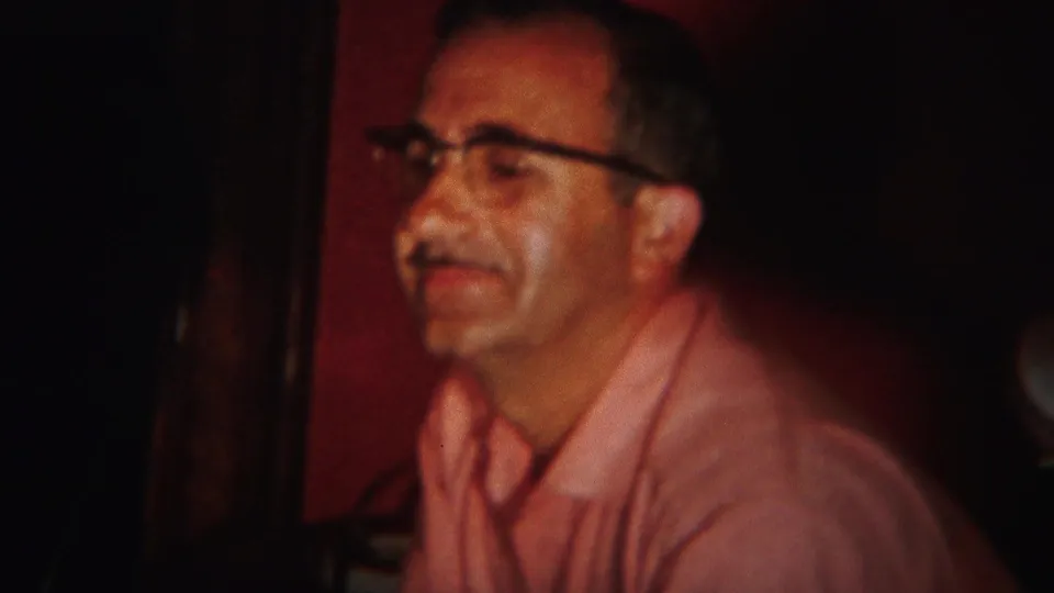 In the 1951 Westville interior, a man in a pink shirt and glasses sits in warm light as if speaking or listening.