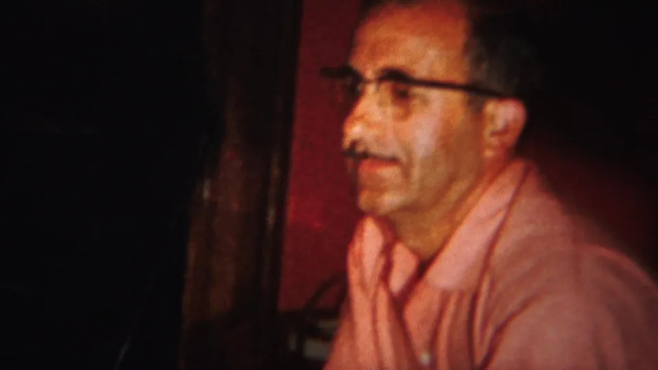 A man in glasses and a pink shirt sits in a dark Westville, New Jersey interior, with warm 8mm color and soft focus.