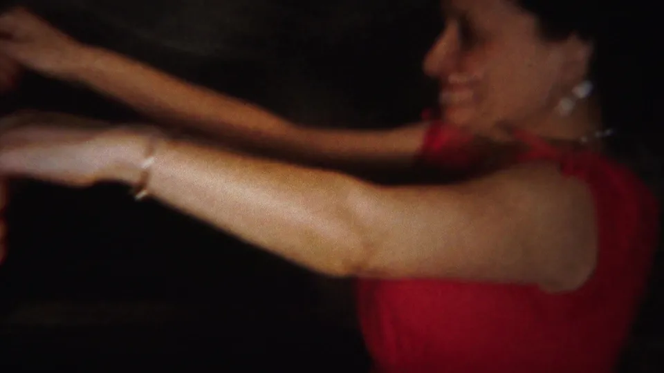 A smiling young woman in a red sleeveless dress and pearl-like earrings extends her arm while dancing in the 1951 home movie.