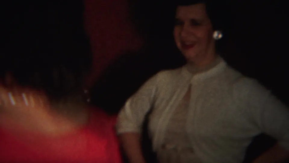 A red-dressed woman crosses the foreground as a smiling woman in a light blouse watches at the 1951 indoor party.