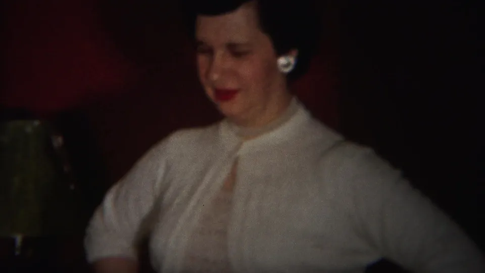 The woman in a light dress looks downward near a red wall and small lamp in the 1951 Westville party footage.