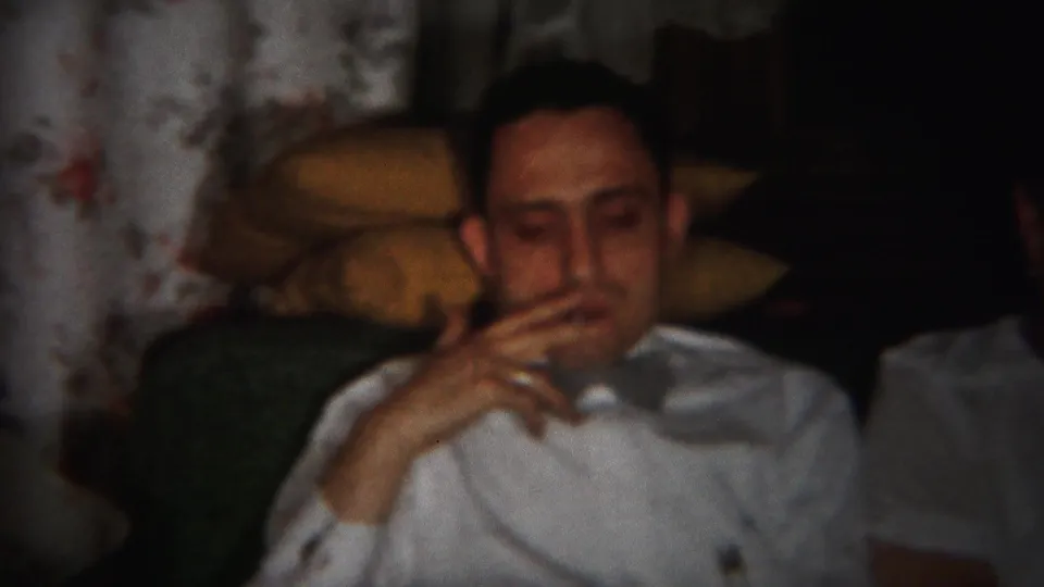 1951 Westville home-movie frame showing a seated man in a white shirt and bow tie holding a cigarette on a floral couch.
