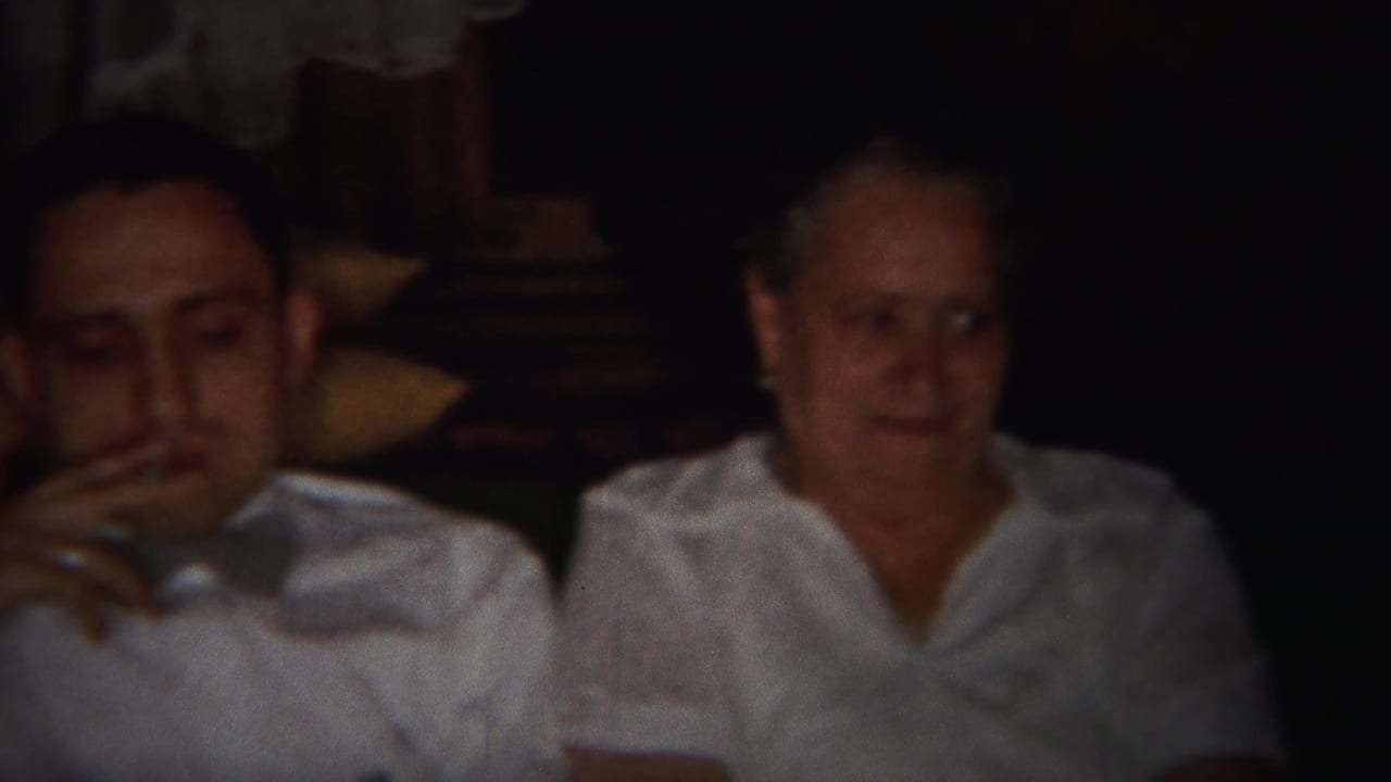 Man in a white shirt holds a cigarette beside an older woman on a couch.
