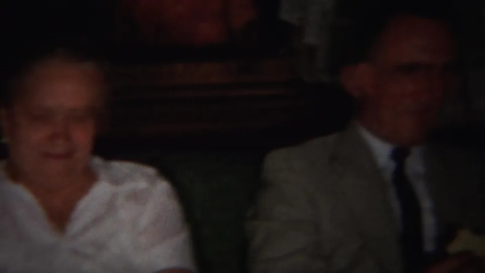 1951 Westville home-movie frame showing an older woman and a man in a suit seated together in a dim interior.
