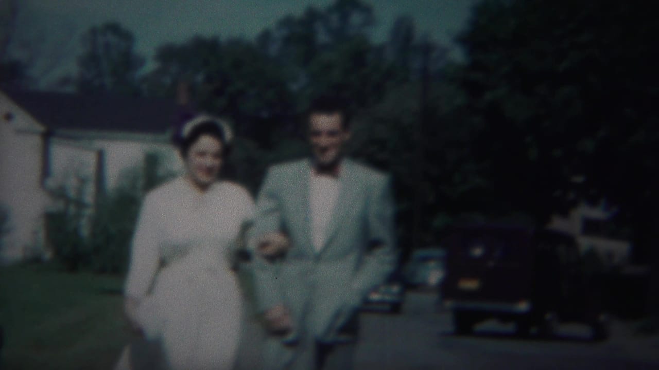 Couple walking arm in arm on a sidewalk with parked cars, lawn, trees, and houses nearby.