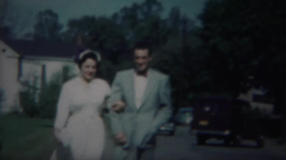 A couple walks arm in arm on a suburban sidewalk in Westville, New Jersey, with 1950s cars and houses behind them.