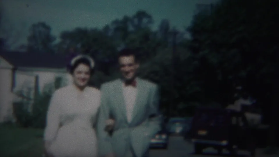 The couple continues walking toward the camera on a Westville sidewalk, smiling amid houses, trees, and parked cars.