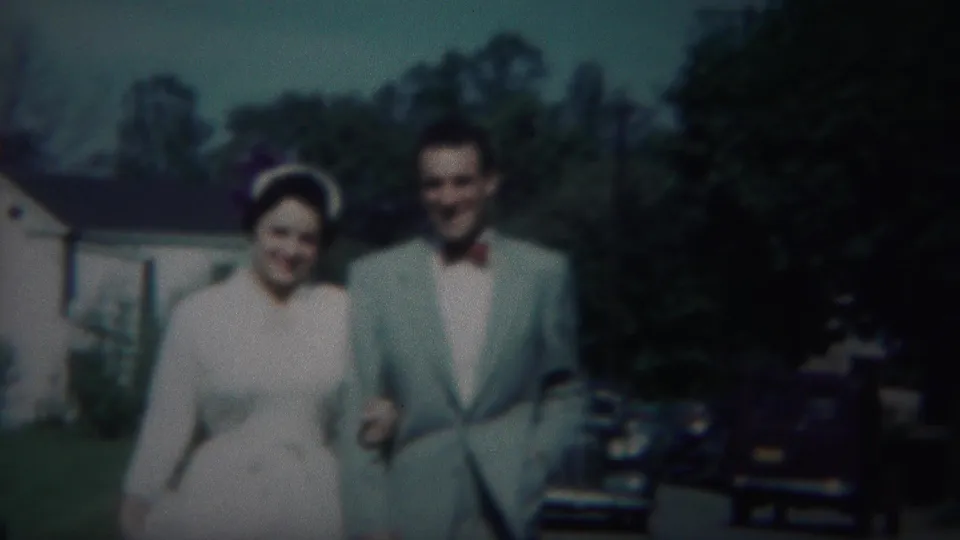 A man in a light suit and a woman in a light dress walk arm in arm on a 1951 Westville suburban sidewalk.