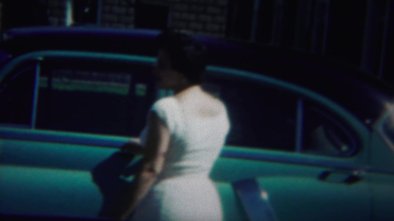 Woman in a white dress seen from behind beside a blue car outside a brick house.