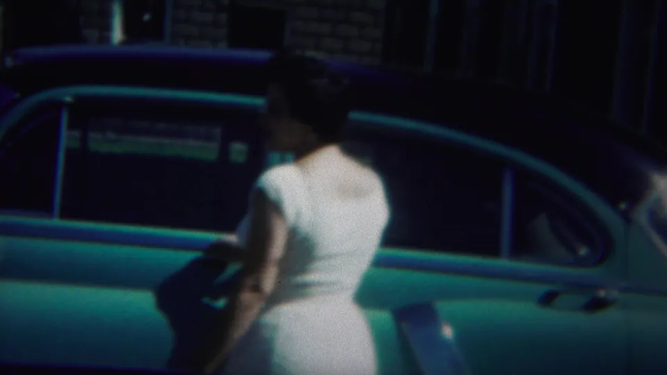 In Westville, New Jersey, a woman in a white dress stands beside a rounded blue car outside a brick residential building.