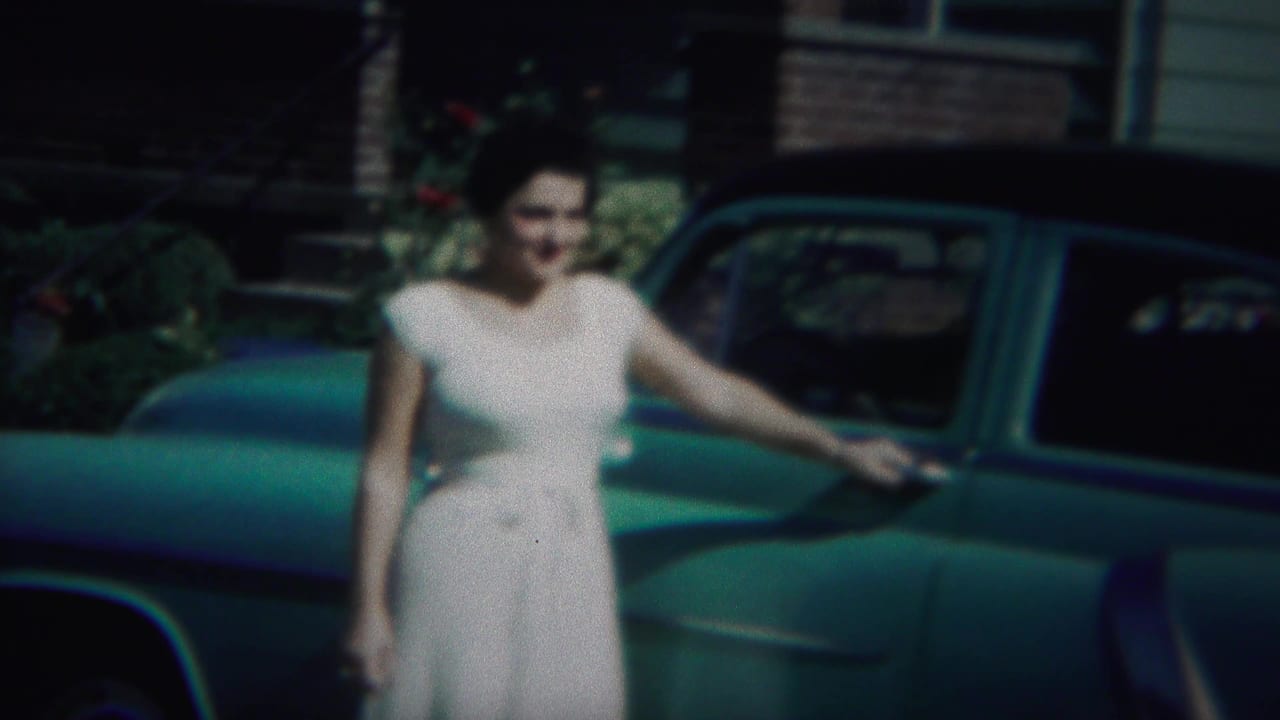 Woman in a white dress faces the camera with one hand near a blue car.