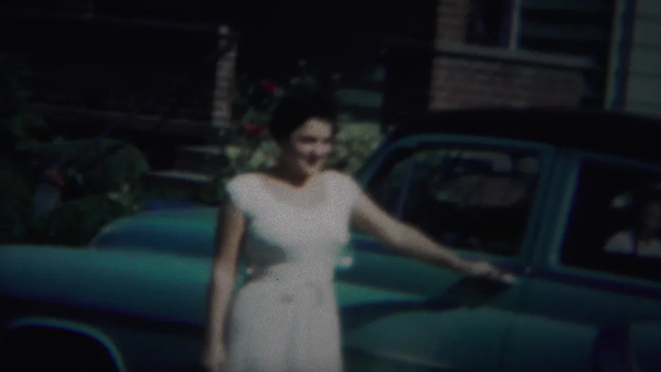 A smiling woman in a white dress poses near the blue car, with dark windows and brickwork visible in the background.