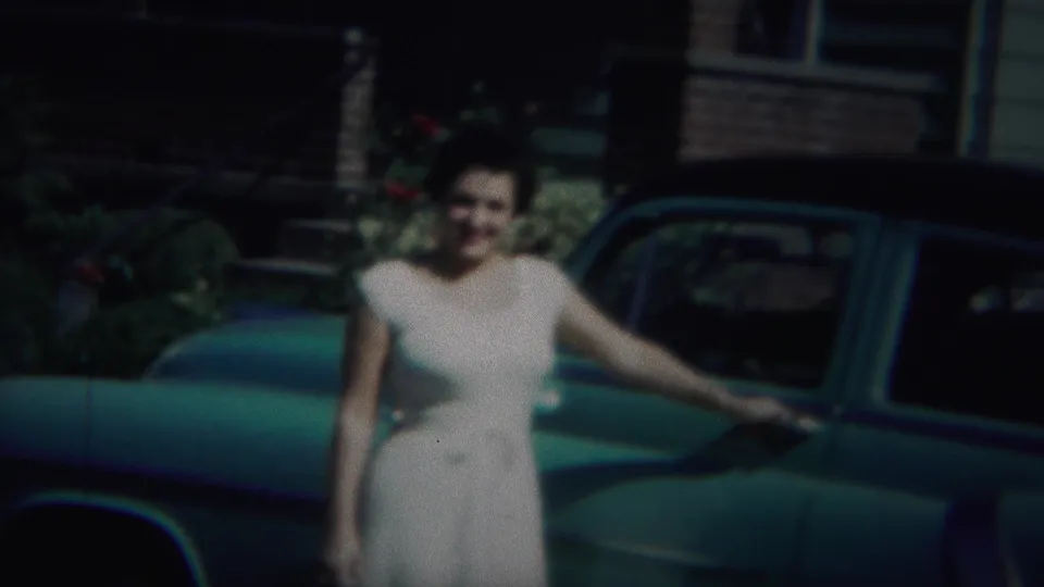 The woman continues smiling beside the blue car, her arm extended toward the door area as the house remains in shadow behind her.