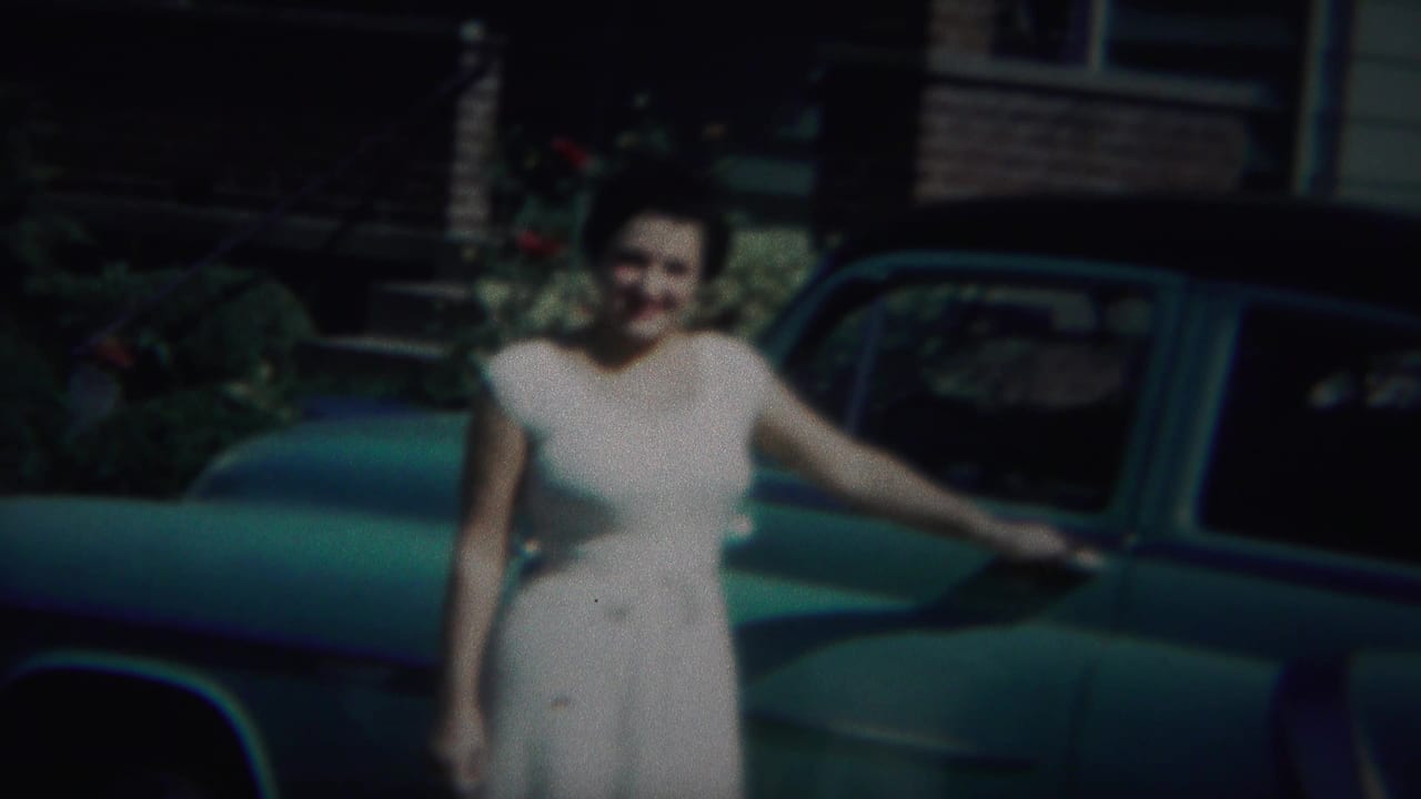 Woman in a white dress stands beside a green vintage car outside a house.