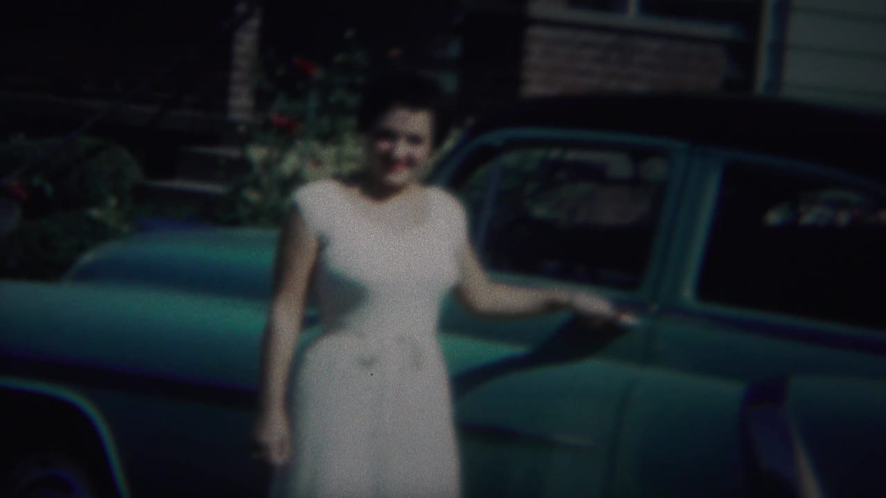 The woman remains posed beside the green car, with a house facade and greenery visible behind her in the 1951 home movie frame.