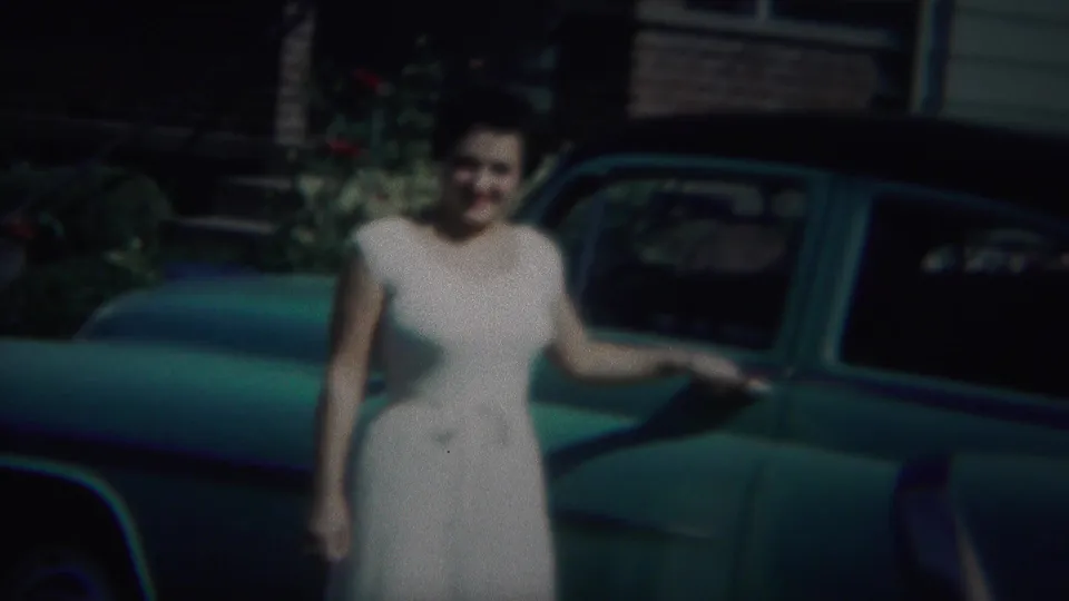 The woman remains posed beside the green car, with a house facade and greenery visible behind her in the 1951 home movie frame.