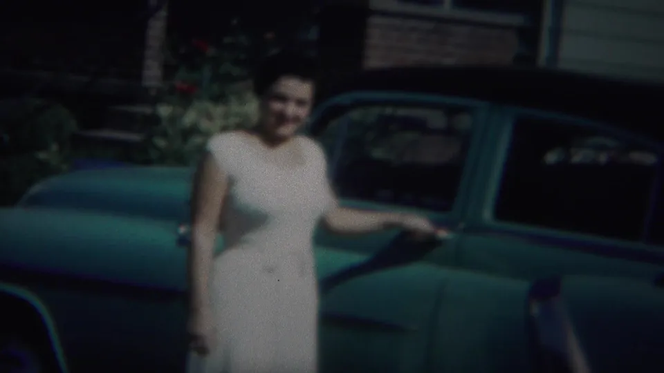 A closer 8mm frame shows the woman in a white dress beside the green car, with residential siding, brickwork, and plants behind her.