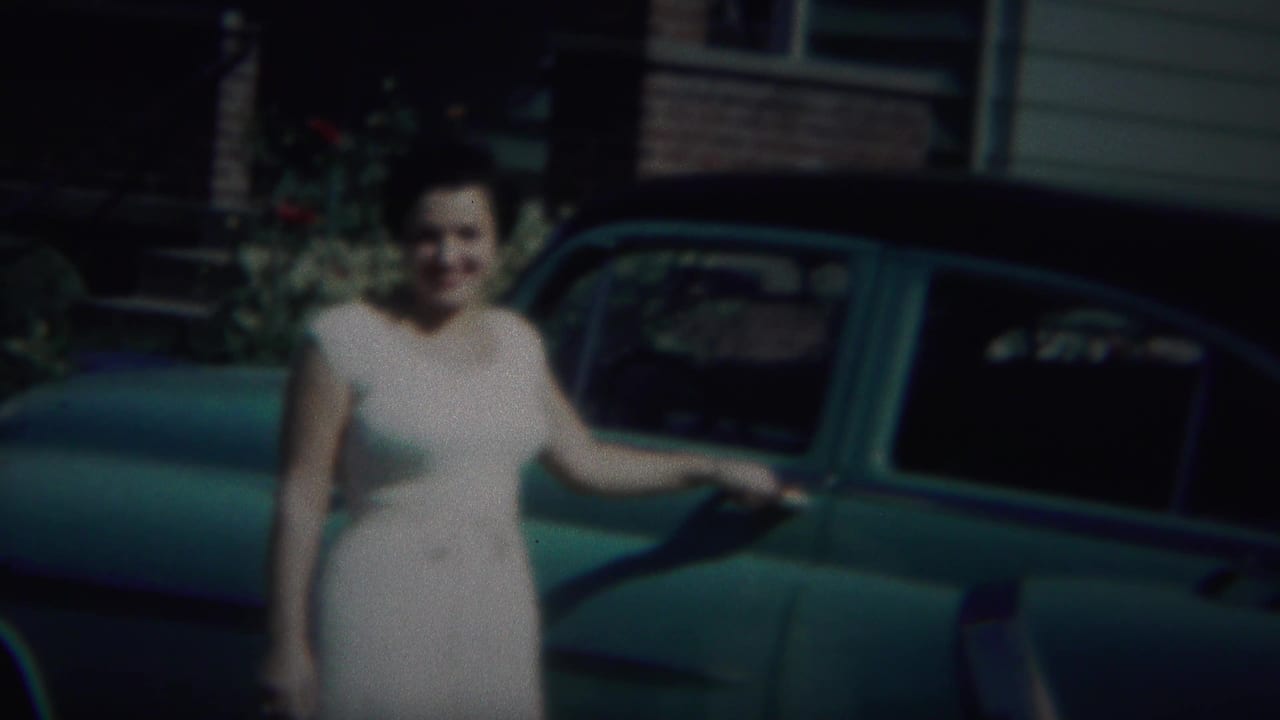 Woman in a light dress stands beside a teal vintage car near a house.