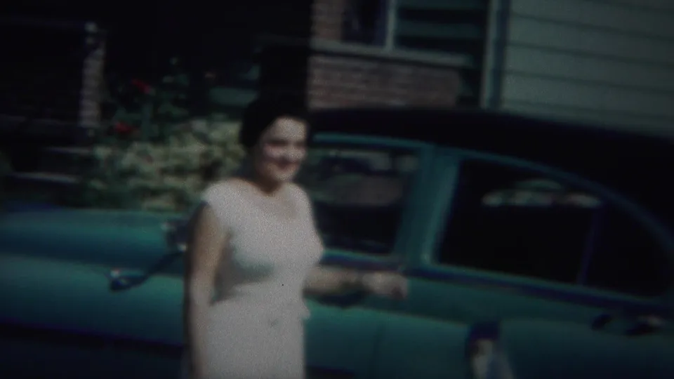 The camera holds on the woman beside the teal car, with the house and garden details soft in the background.