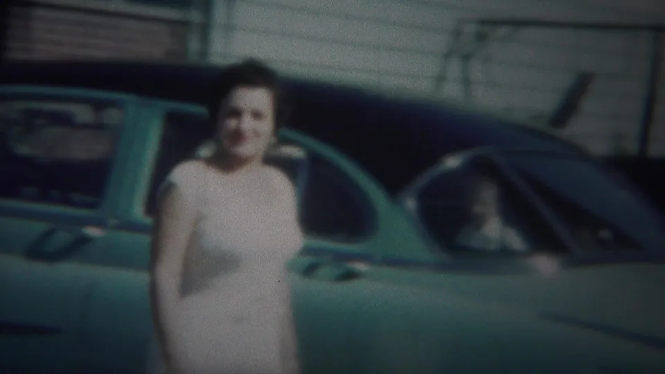 The woman stands near the teal car while another person appears seated inside, in a 1951 Westville home-movie frame.