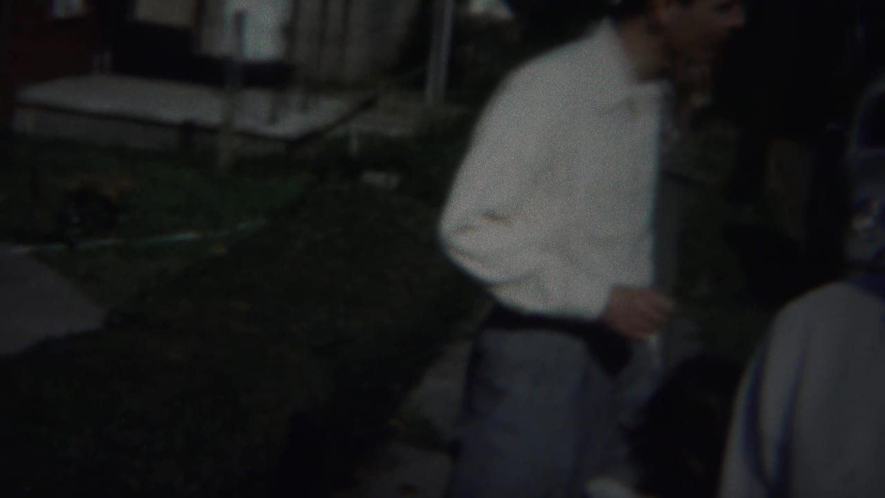 Man in a white shirt stands outdoors near a lawn and house, with another figure partly visible.