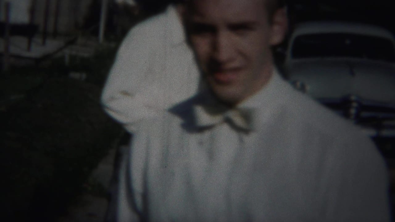 Man in a white shirt and bow tie faces the camera with a parked car behind him.